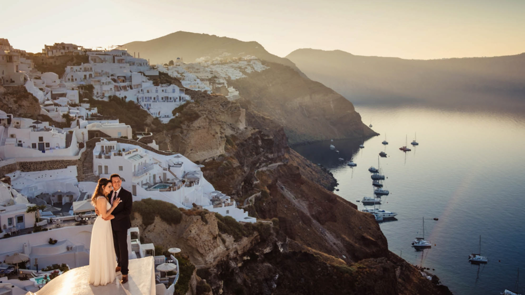 Para młoda na tle wybrzeża Santorini. Białe domki i czarne wulkaniczne skały.