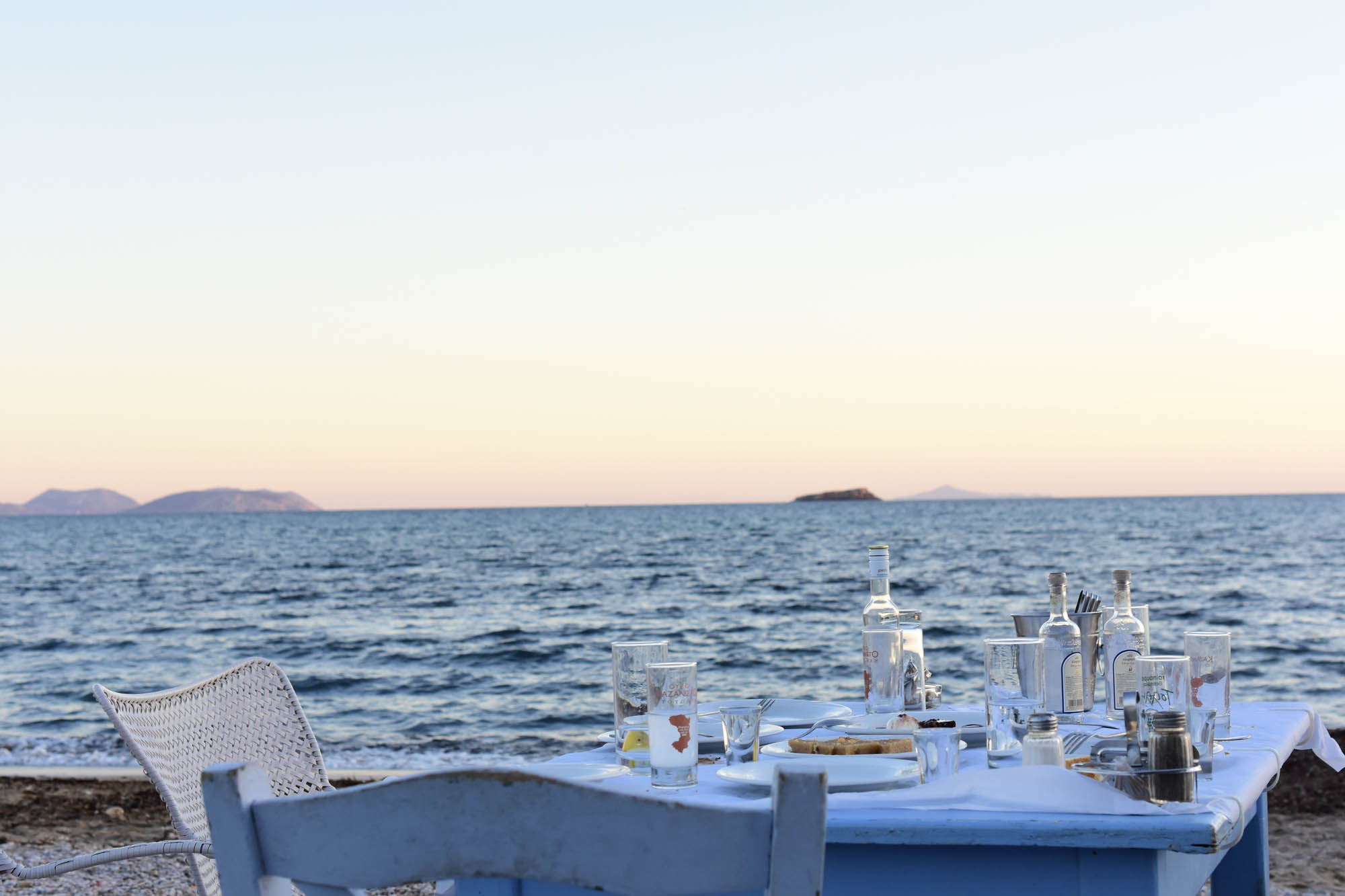 stół nad brzegiem mora a na nim trunki i szklanki, wino i ouzo