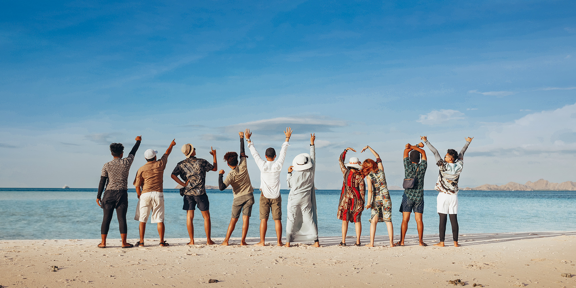 ludzie stojący na brzegu morza w kolorowych ubraniach i podnoszący ręce do góry z radości