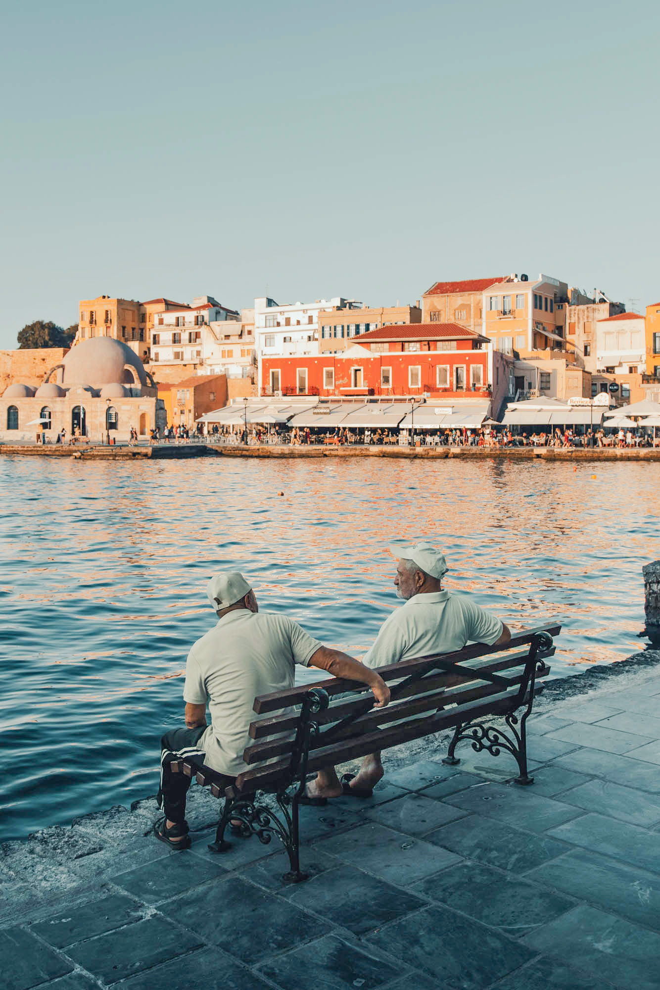 uroczy widok na port w chanii, dwóch greków siedzi na ławce i rozmawia a w tle morze