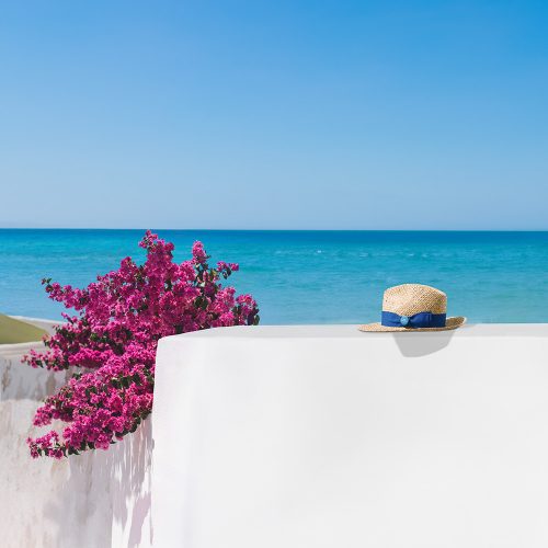 widok na morze zza muru porośniętego różowymi kwiatami, biały mur, niebieskie morze, kwiaty, typowy obrazek dla grecji