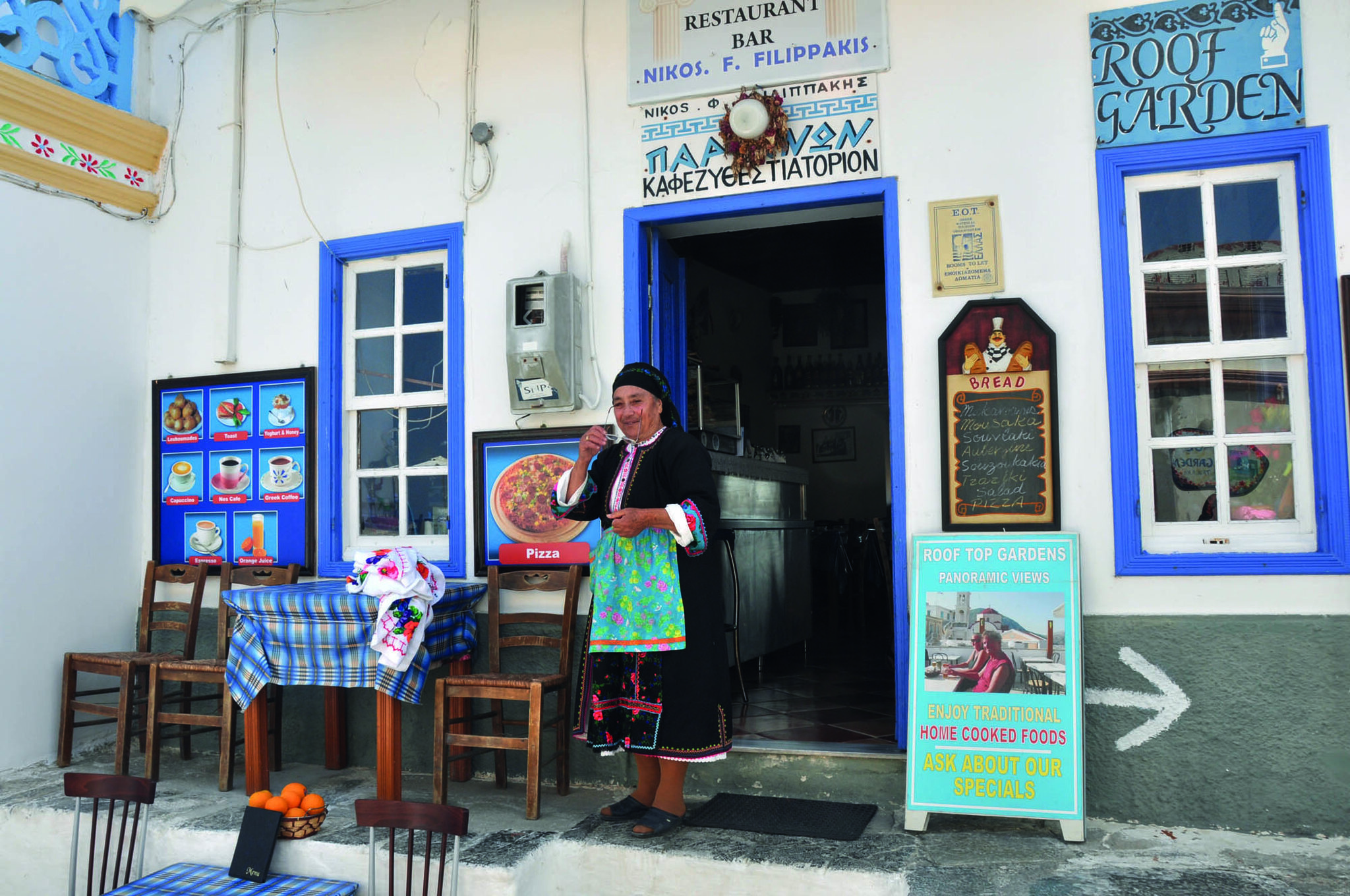 typowa grecka kobieta - starsza pani przed sklepem, miejscem w grecji z jedzeniuem, tawerną i stolikami, niebieskie okna