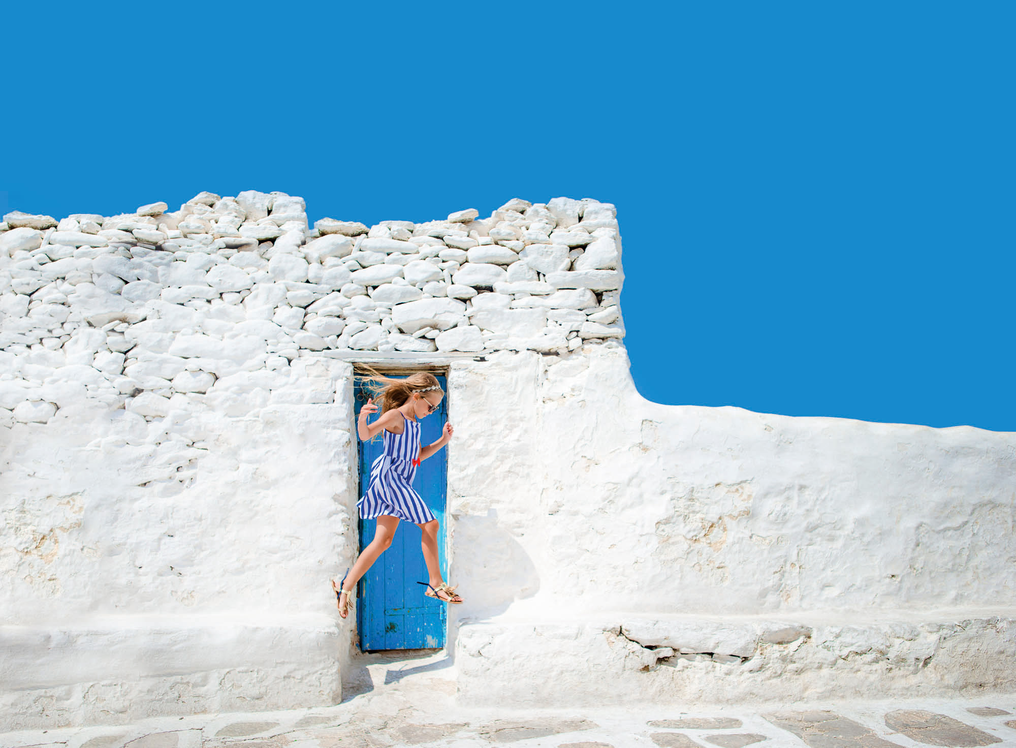niebieskie niebo i biała ściana, typowy obrazek w grecji, na tle ściany skacze dziewczynka w niebieskiej sukience, idylliczny widok