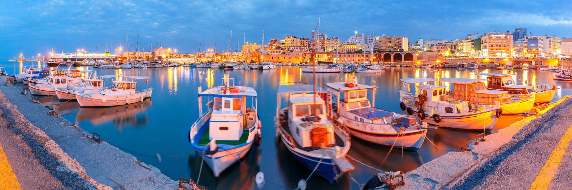 widok na posrt wenecki w heraklionie oświetlony światłami wielkiego miasta, oraz łódki stojące w marinie, wieczorna panorama