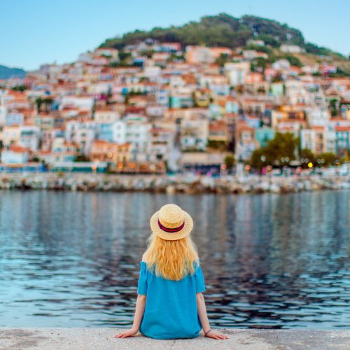 grecja, dziewczyna patrzy na oddalone od brzegu maisteczko i morze, dziewczyna w kapeluszy na brzegu