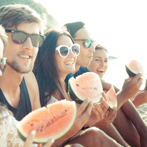 grupa ludzi zajada arbuza na plaży, grupa przyjaciół, ludzie